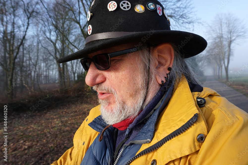 Senior man with sunglasses, a western hat with pins, black earrings and a yellow raincoat is standing in a winter landscape Senior man with sunglasses, a western hat with pins, black earrings and a yellow raincoat is standing in a winter landscape