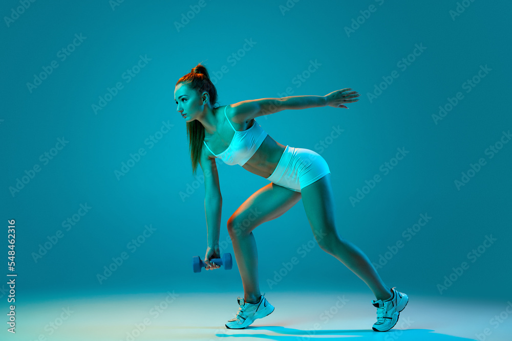 Young sportive girl in white sportswear training with dumbbells isolated over blue studio background in neon light. Keeping balance
