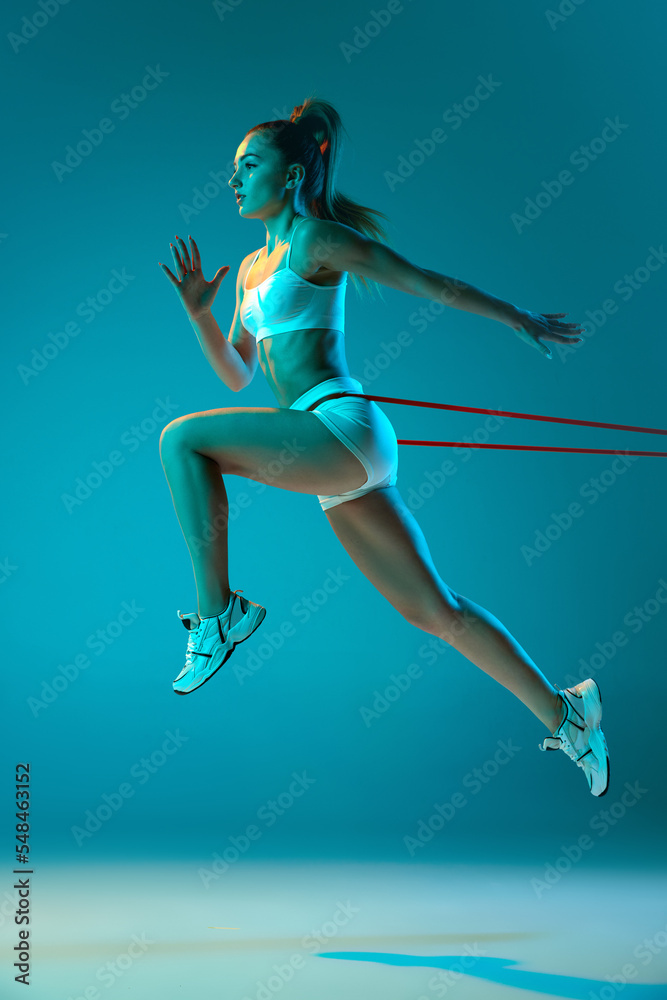 Young sportive girl in white sportswear training with sports expanders isolated over blue studio background in neon light. Run