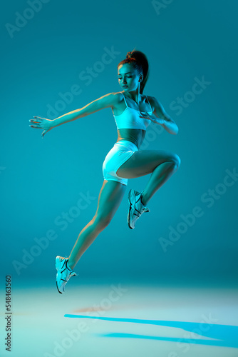 Young sportive girl in white sportswear training, jumping in a run isolated over blue studio background in neon light