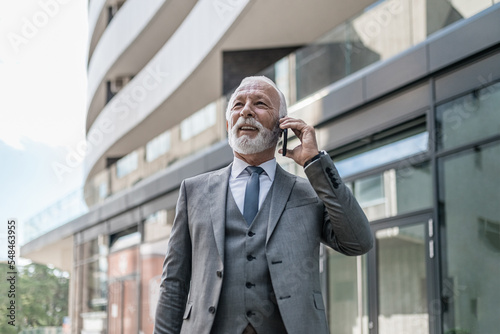 Smiling businessman talking on smart phone while standing outside the office building.