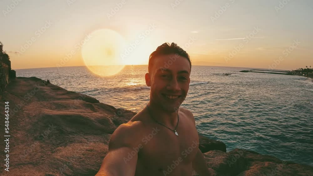 One happy and cheerful young man looking at the camera and walking at the beach alone with sunset at the background. Male person having fun and enjoying freedom taking selfie.