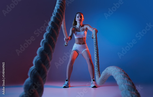 Young sportive girl training with sports equipment, ropes isolated over gradient blue purple studio background in neon light. Power