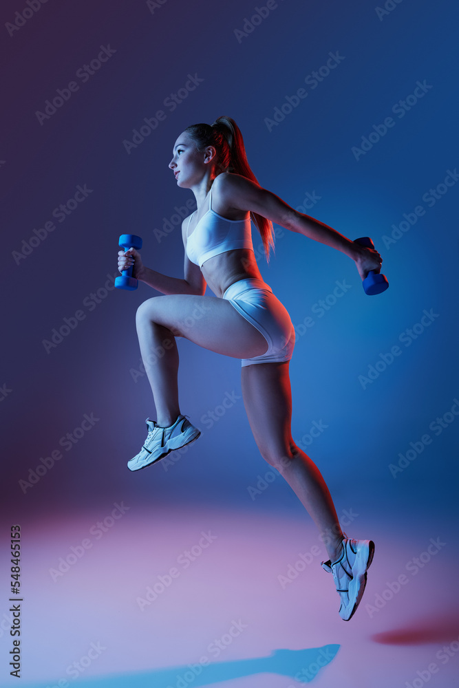 Young sportive girl in white sportswear training with dumbbells isolated over gradient blue purple studio background in neon light. High jump