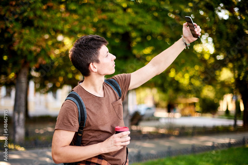 hipster man in brown T-shirt walking and take selfie on phone camera in summer green city park