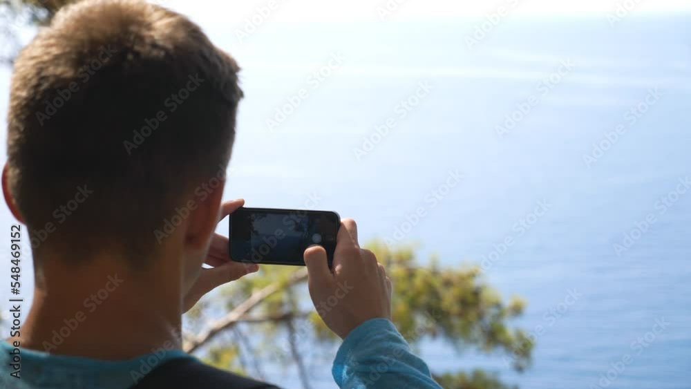 Unrecognizable man taking photo of scenic seascape on his smartphone. Young guy using phone to get beautiful pictures of sea from hill top. Male hiker enjoying travel during summer vacation. Slow mo Unrecognizable man taking photo of scenic seascape on his smartphone. Young guy using phone to get beautiful pictures of sea from hill top. Male hiker enjoying travel during summer vacation. Slow mo
