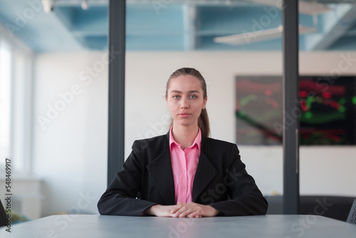 Successful young female leader in a suit with a pink shirt sitting in a modern glass office with a determined smile.