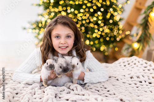 Happy little girl with three rabbits symbols of the new year 2022 embraces animals on the background of a Christmas tree with blurred lights