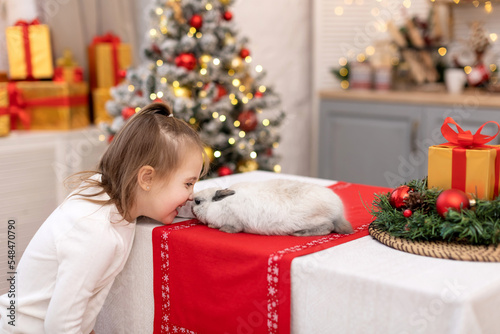 A happy little girl laughs and rubs her noses with a little rabbit on the background of a Christmas tree with blurred lights