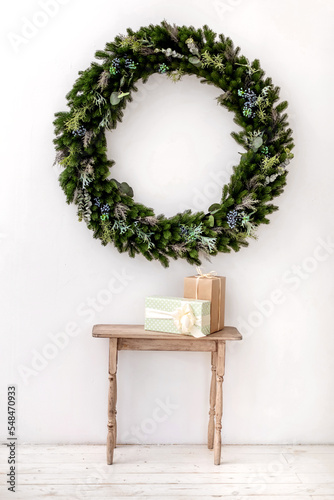 Minimalistic Christmas card with a wreath of branches on a white wall background, wooden table with gifts. Copy space for text