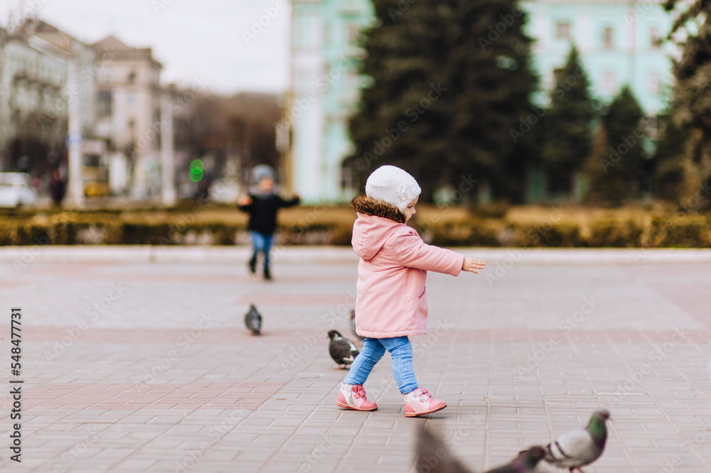 A little beautiful girl, a child in a pink jacket, walks down the street in the city, feeding pigeons. A little beautiful girl, a child in a pink jacket, walks down the street in the city, feeding pigeons.