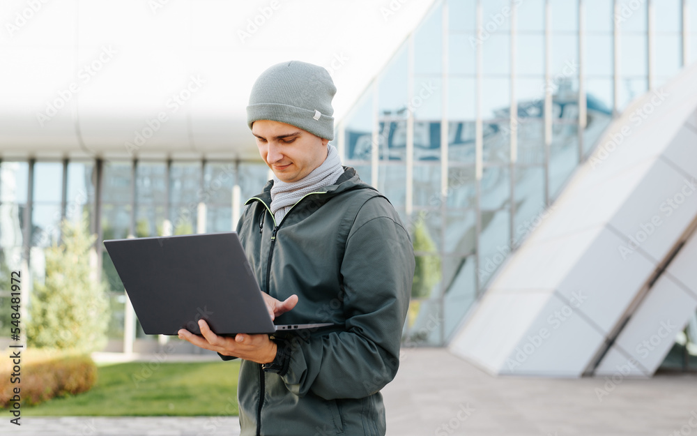Young man using laptop while working outdoors. Student or freelancer in outerwear typing on laptop keyboard while standing against backdrop of university or business office Young man using laptop while working outdoors. Student or freelancer in outerwear typing on laptop keyboard while standing against backdrop of university or business office