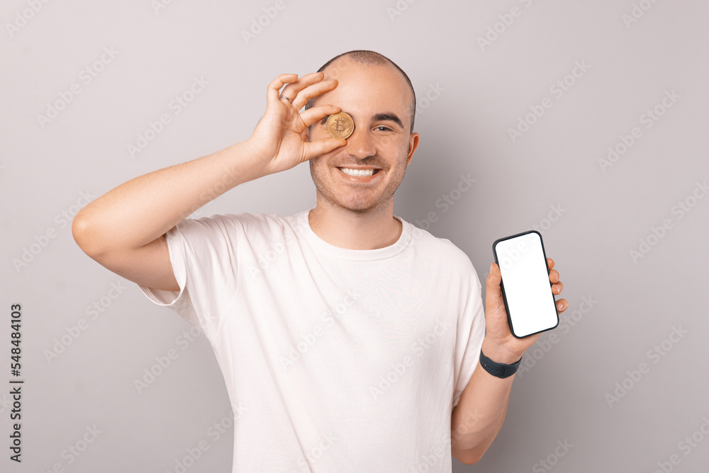 Cheerful young man in white t-shirt showing bitcoin and blank white screen on smartphone Cheerful young man in white t-shirt showing bitcoin and blank white screen on smartphone