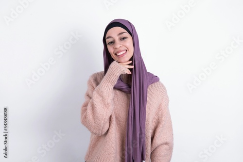 Optimistic muslim woman wearing hijab and knitted sweater over white background keeps hands partly crossed and hand under chin, looks at camera with pleasure.