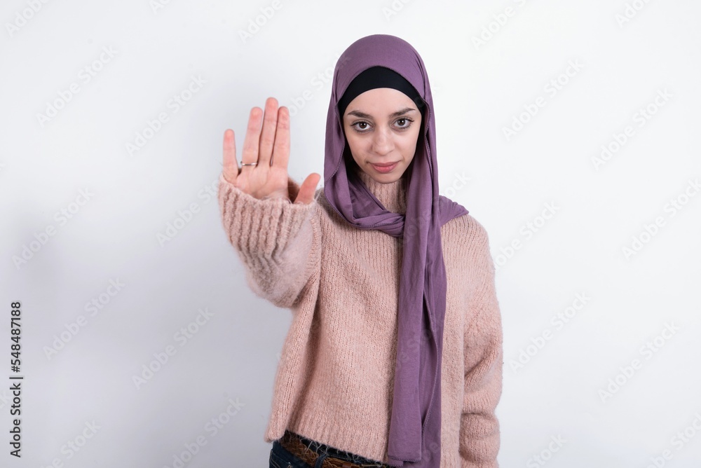 beautiful muslim woman wearing hijab and warm jumper over white background doing stop sing with palm of the hand. Warning expression with negative and serious gesture on the face. beautiful muslim woman wearing hijab and warm jumper over white background doing stop sing with palm of the hand. Warning expression with negative and serious gesture on the face.