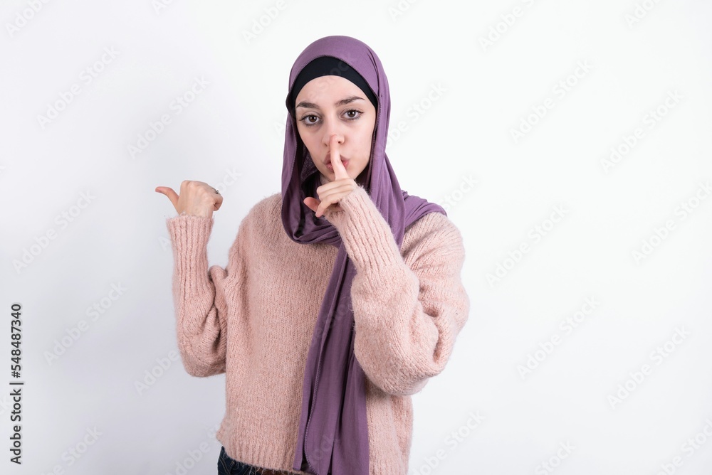 Φωτογραφία beautiful muslim woman wearing hijab and warm jumper over white background asking to be quiet with finger on lips pointing with hand to the side