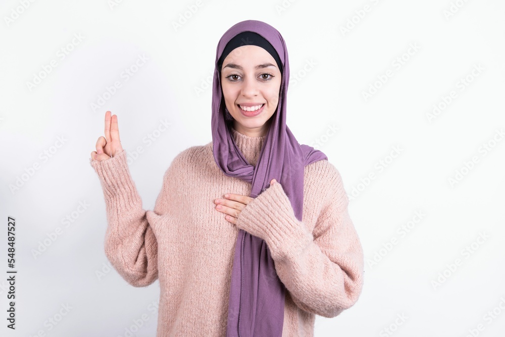 beautiful muslim woman wearing hijab and warm jumper over white background smiling swearing with hand on chest and fingers up, making a loyalty promise oath. beautiful muslim woman wearing hijab and warm jumper over white background smiling swearing with hand on chest and fingers up, making a loyalty promise oath.