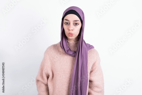 Shot of pleasant looking beautiful muslim woman wearing hijab and warm jumper over white background , pouts lips, looks at camera, Human facial expressions