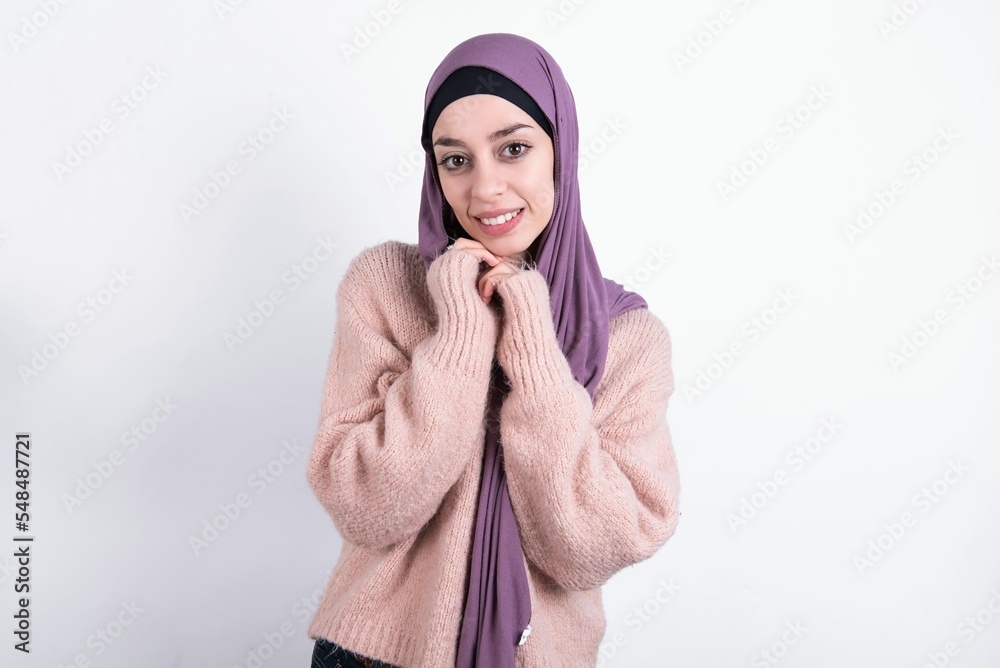 Positive beautiful muslim woman wearing hijab and warm jumper over white background smiles happily, glad to receive pleasant news from interlocutor, keeps hands together. People emotions concept. Positive beautiful muslim woman wearing hijab and warm jumper over white background smiles happily, glad to receive pleasant news from interlocutor, keeps hands together. People emotions concept.