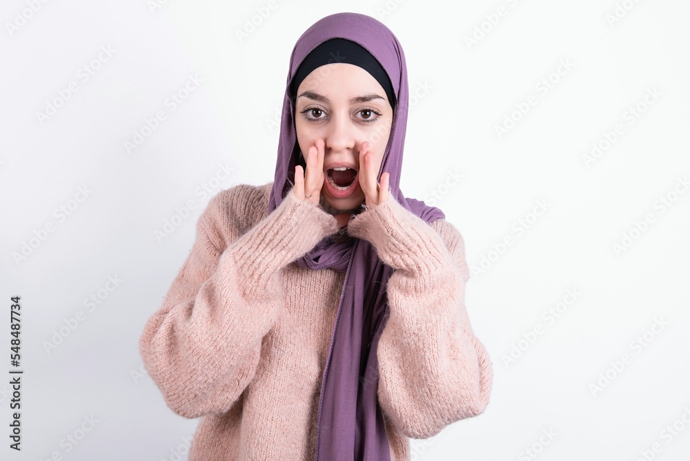 Φωτογραφία beautiful muslim woman wearing hijab and warm jumper over white background shouting excited to front