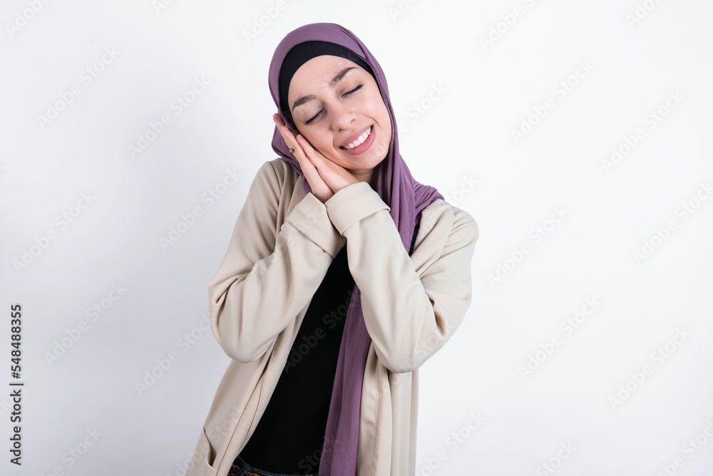 young beautiful muslim woman wearing hijab and jacket over white background leans on pressed palms closes eyes and has pleasant smile dreams about something young beautiful muslim woman wearing hijab and jacket over white background leans on pressed palms closes eyes and has pleasant smile dreams about something