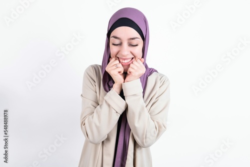 Cheerful young beautiful muslim woman wearing hijab and jacket over white background has shy satisfied expression, smiles broadly, shows white teeth, People emotions