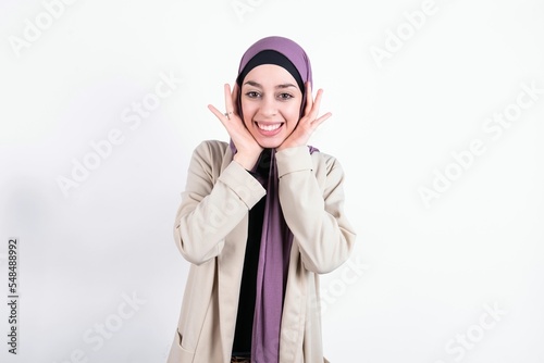 Happy young beautiful muslim woman wearing hijab and jacket over white background touches both cheeks gently, has tender smile, shows white teeth, gazes positively straightly at camera,