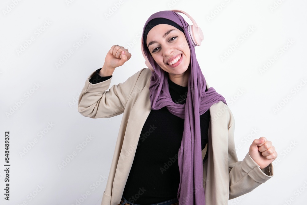 Carefree young beautiful muslim woman wearing hijab and jacket over white background with toothy smile raises arms dances carefree moves with rhythm of music listens music from playlist via headphones Carefree young beautiful muslim woman wearing hijab and jacket over white background with toothy smile raises arms dances carefree moves with rhythm of music listens music from playlist via headphones