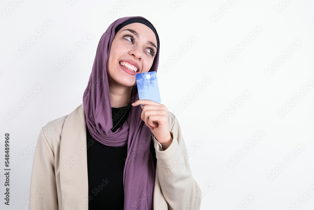 Lovely young beautiful muslim woman wearing hijab and jacket over white background showing credit card and looking away at copy space. Pensive Lovely young beautiful muslim woman wearing hijab and jacket over white background showing credit card and looking away at copy space. Pensive
