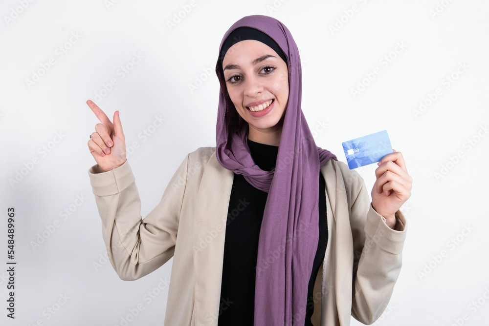 Smiling young beautiful muslim woman wearing hijab and jacket over white background showing debit card pointing finger empty space Smiling young beautiful muslim woman wearing hijab and jacket over white background showing debit card pointing finger empty space