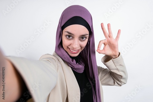 Portrait of pretty cheerful young beautiful muslim woman wearing hijab and jacket over white background make selfie show okey symbol