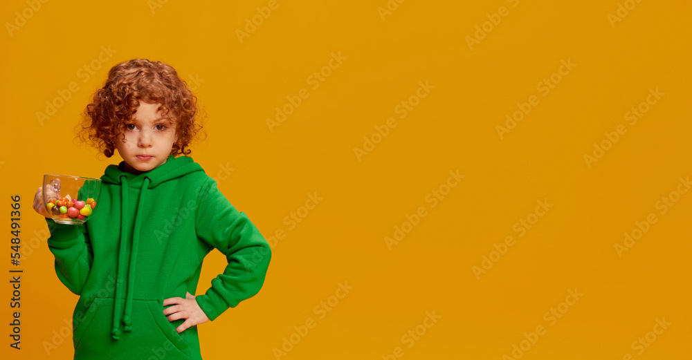 Portrait of cute little girl, child with curly red hair posing with candies in cup isolated over yellow background. Flyer