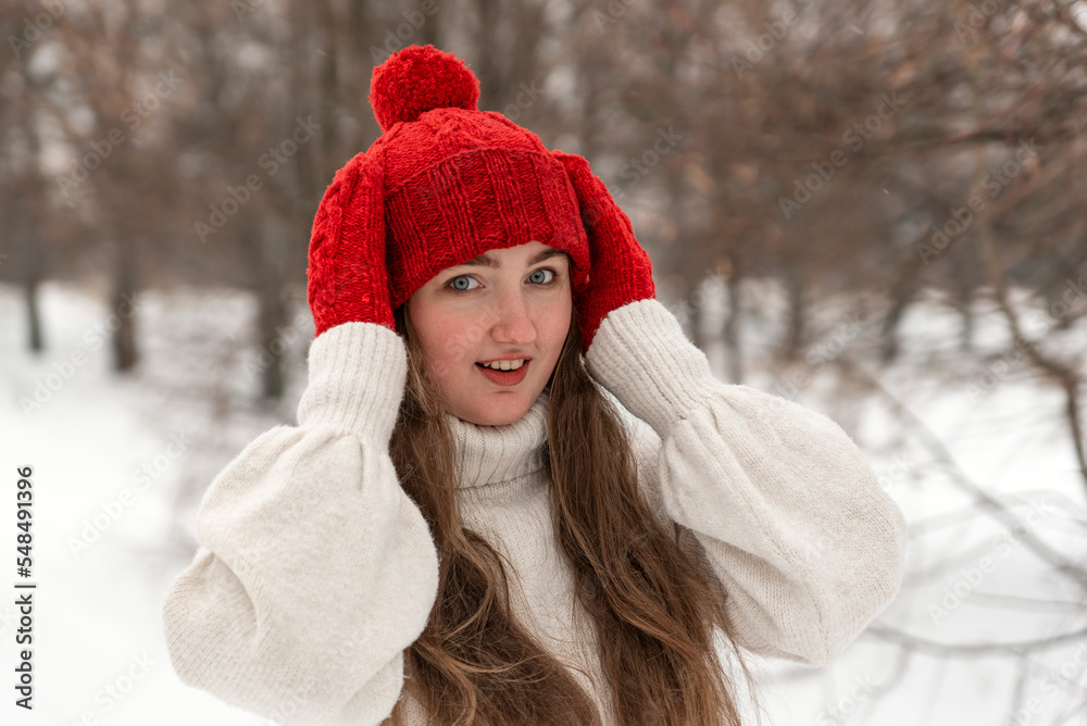 Happy blue-eyed girl in knitted red hat and mittens and woolen sweater in winter forest. Female knitwear Happy blue-eyed girl in knitted red hat and mittens and woolen sweater in winter forest. Female knitwear