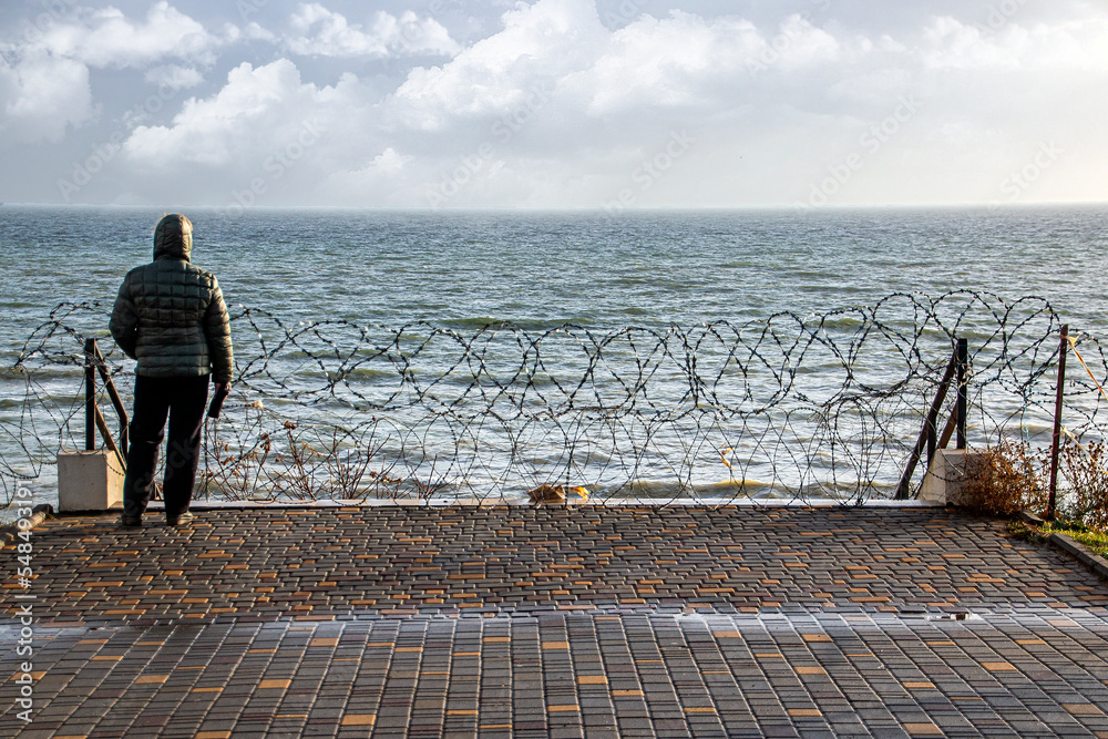 A woman sadly looks at the sea horizon through barbed wire during the war in Ukraine