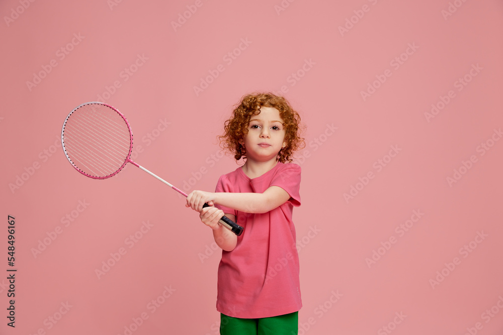 Portrait of cute little girl, child with curly red hair playing badminton isolated over pink background Portrait of cute little girl, child with curly red hair playing badminton isolated over pink background