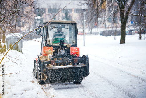 Tractor with brush clear sidewalk from snow and ice during blizzard. Tractor with snowplow and rotary brush cleaning pedestrian road from snow in winter. Tractor mounted sweeper brush and snow plow