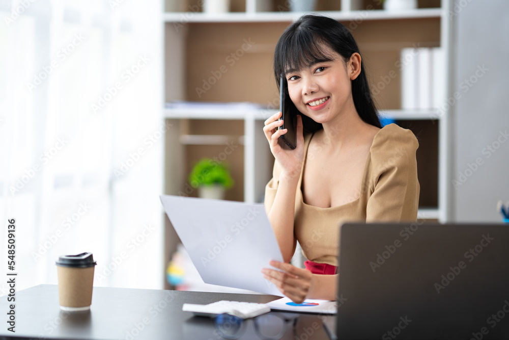 Happy young asian woman talking on the mobile phone and smiling while sitting at her working place in office. Happy young asian woman talking on the mobile phone and smiling while sitting at her working place in office.