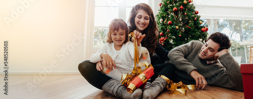 Fotografija Family opening gifts by the Christmas tree on Christmas morning - kid excited about unwrapping a present from her parents
