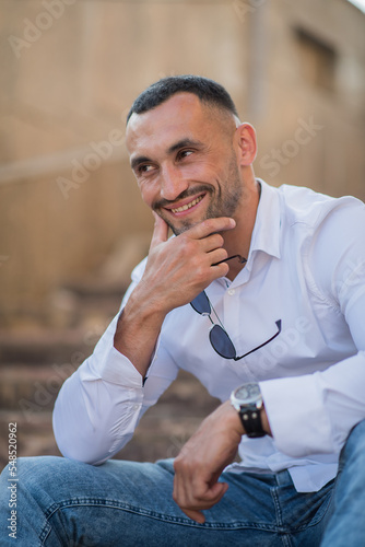 Portrait of a handsome confident stylish man. Sexy modern man wearing a white shirt. Fashionable man posing against the backdrop of a street in the city at sunset, holding sunglasses in his hands