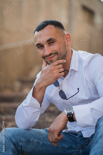 Handsome stylish man. Sexy modern man wearing a white shirt. Fashionable man posing against the backdrop of a street in the city at sunset, holding sunglasses in his hands