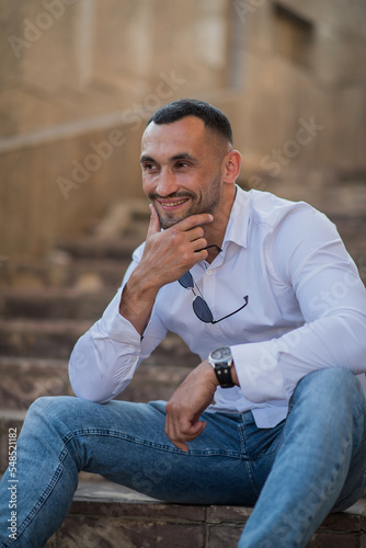 Portrait of a handsome confident stylish man. Sexy modern man wearing a white shirt. Fashionable man posing against the backdrop of a street in the city at sunset, holding sunglasses in his hands