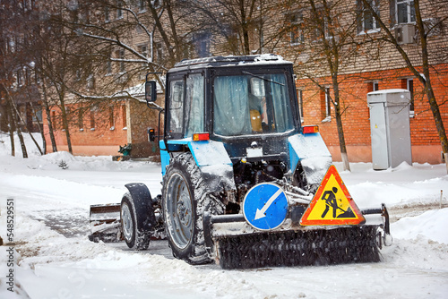 Tractor with brush and scoop cleaning snow at city street during heavy snowfall at winter day. Tractor clearing snowy streets with snow plow and ratary brush. Vehicle sweeps snow on pedestrian road