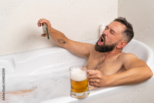 Bearded man watching football on smartphone while drinking a beer in bath with soapsuds at home