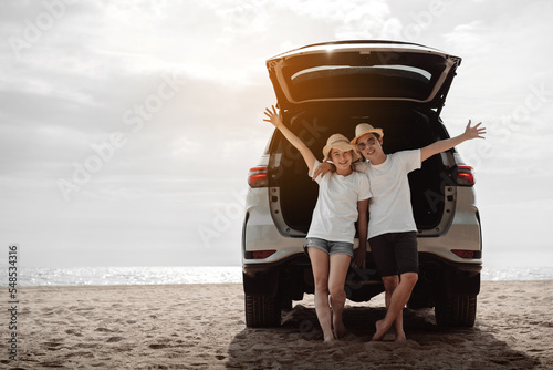 Car road trip travel of couple enjoying beach relaxing on hood of sports utility car. Happy Asian woman, man friends smiling together on vacation weekend holidays on the beach.