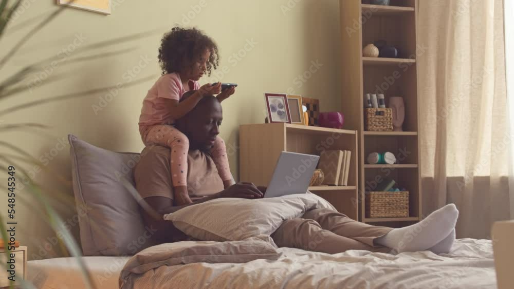 African American man working on laptop in bed while his 3 year old daughter sitting on his shoulders and watching cartoons on smartphone African American man working on laptop in bed while his 3 year old daughter sitting on his shoulders and watching cartoons on smartphone