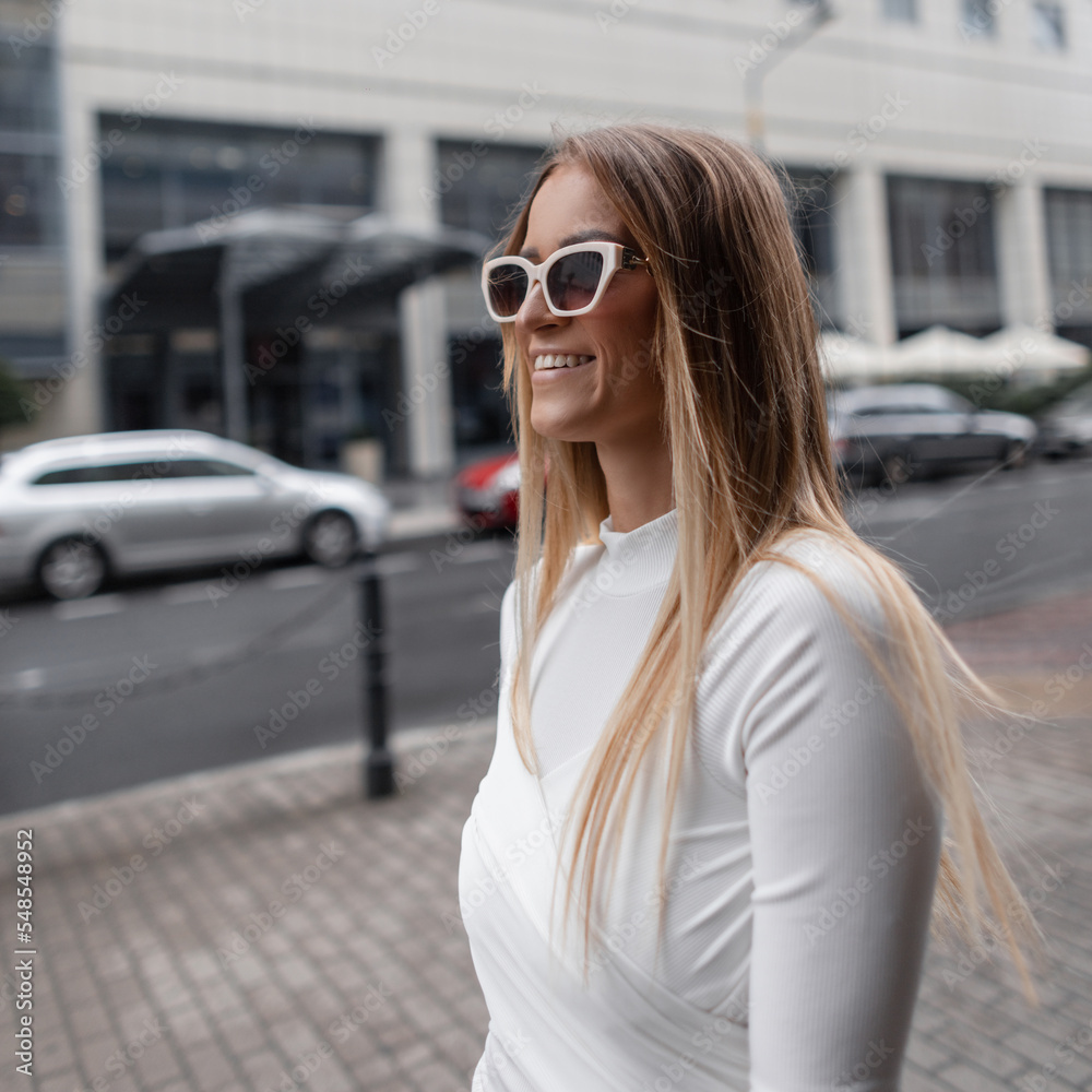 Street female portrait of a beautiful fashion woman with a smile with trendy white sunglasses in a stylish elegant white dress walks in the city