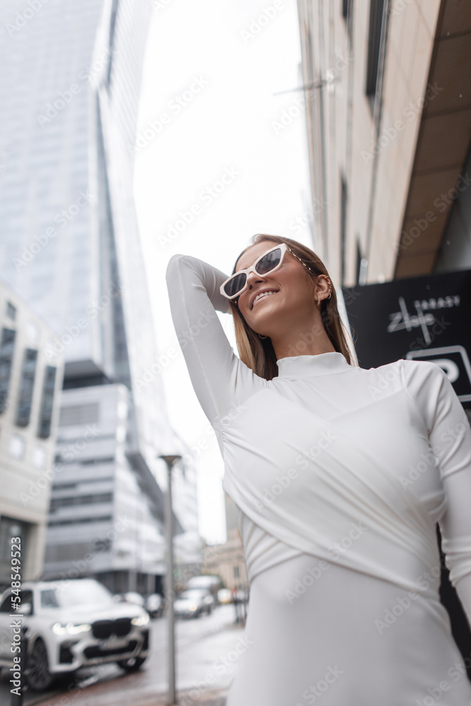 Fashionable beautiful happy vogue girl with white trendy sunglasses in a stylish fashion white dress walks and enjoys in the city. Urban female style look