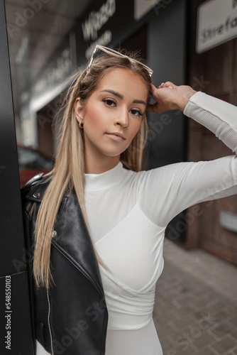 Fashionable pretty girl with beauty face in white fashion dress with leather black jacket walks in the city. urban female rock style clothes © alones
