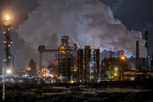 An industrial plant releases a huge cloud of steam at night. A tall mast nearby burns with a bright torch