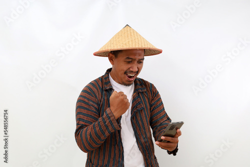 Happy Asian farmer standing while holding a cell phone and clenching his hand. Isolated on white